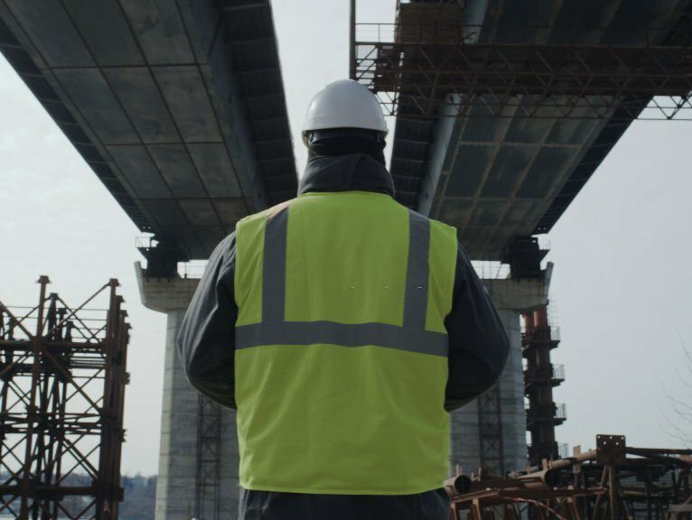 View of anonymous man in uniform and hardhat inspecting unfinished concrete bridge on construction site