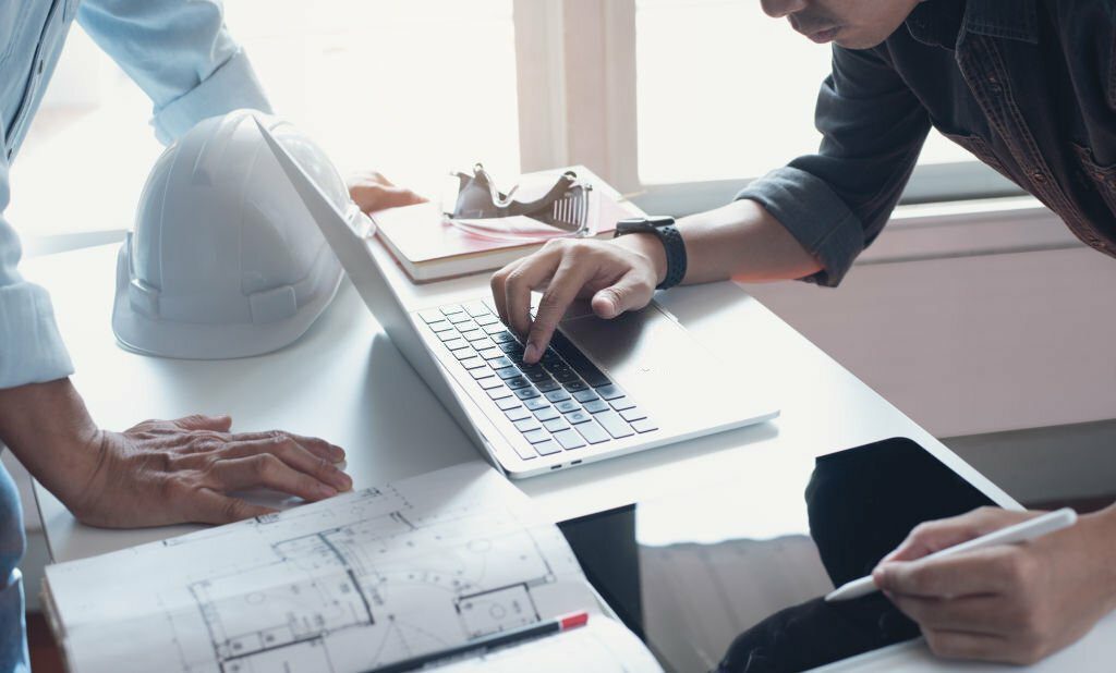 Two  architect colleagues discussing data working on digital tablet and laptop computer with architectural project at construction site on desk at office